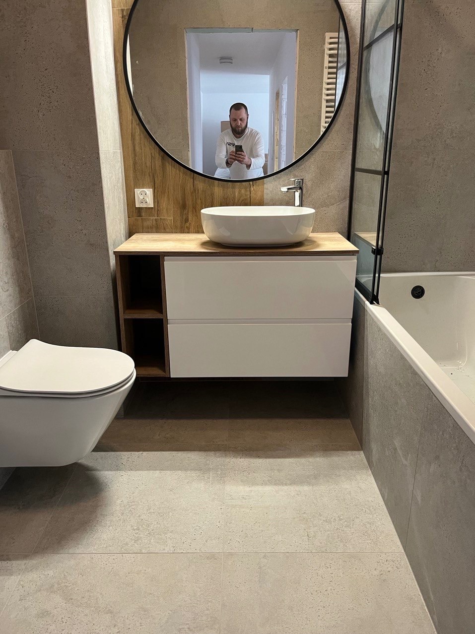 Nowoczesna łazienka z szarą płytką, podwieszaną miską WC i umywalką nablatową w okrągłym lustrze, w którym odbija się osoba robiąca zdjęcie.