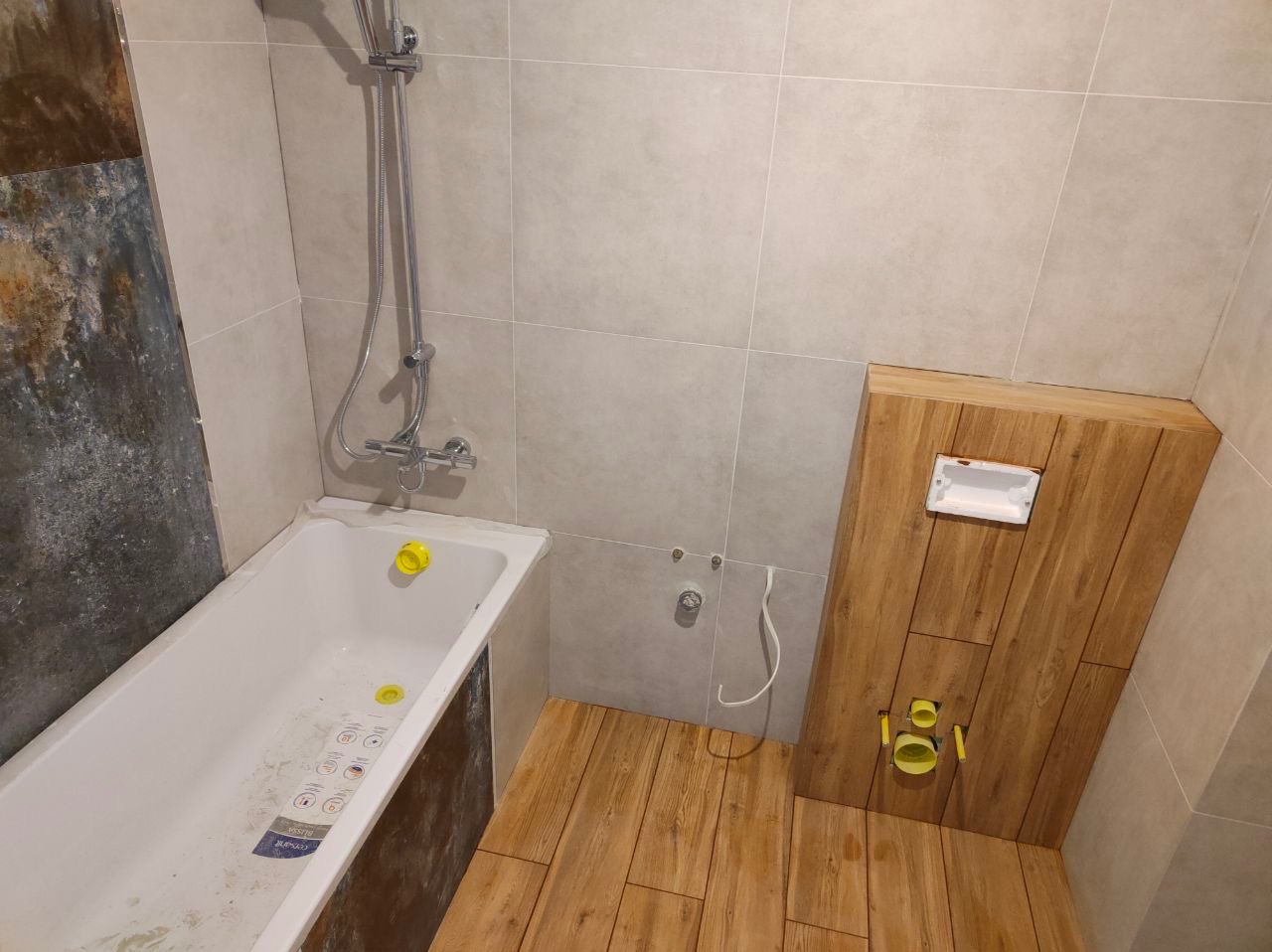 Wnętrze łazienki w trakcie remontu: częściowo wykończona ściana z imitacji drewna, biała wanna z żółtymi zaślepkami, szara glazura, widoczne instalacje wodno-kanalizacyjne.