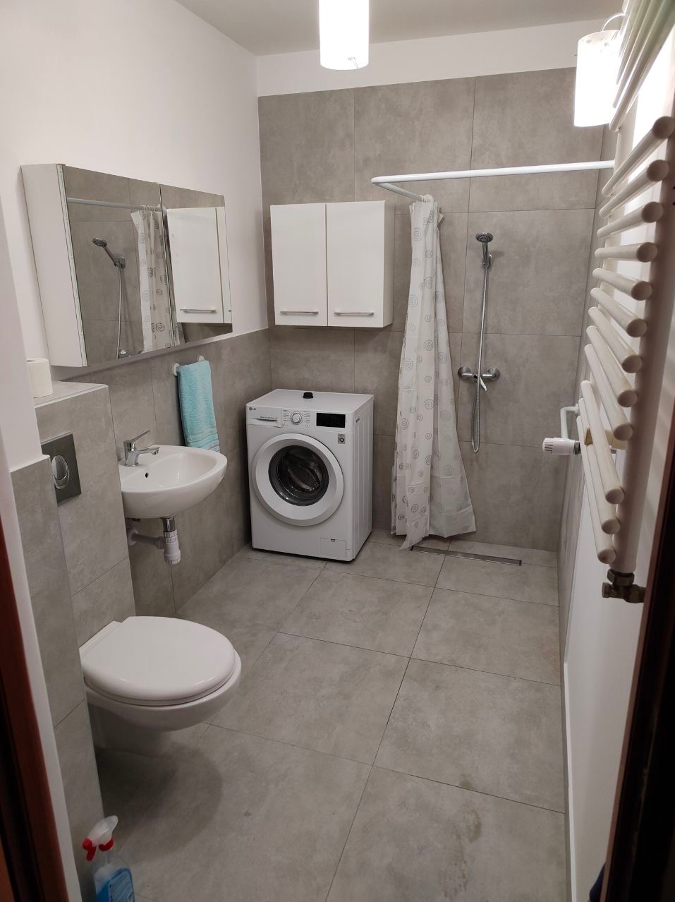 Szara łazienka z pralką LG, umywalką nablatową, szafkami wiszącymi i grzejnikiem drabinkowym, oświetlona dwoma lampami sufitowymi.