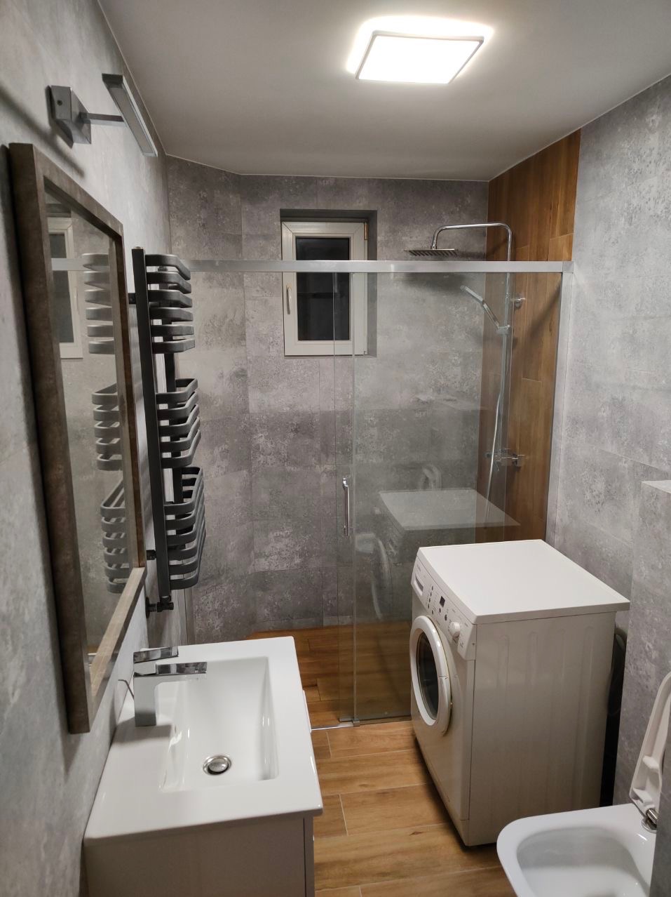 Nowoczesna łazienka z szarymi płytkami imitującymi beton, kabiną prysznicową ze szklaną ścianką, umywalką z szafką, grzejnikiem drabinkowym i pralką w rogu, oświetlona kwadratową lampą sufitową.