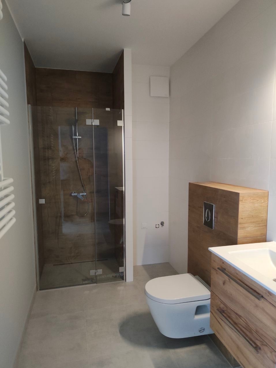 Nowoczesna łazienka z kabiną prysznicową typu walk-in z brązowymi panelami imitującymi drewno, podwieszaną miską WC i szafką z umywalką w kolorze naturalnego drewna z widocznymi słojami.