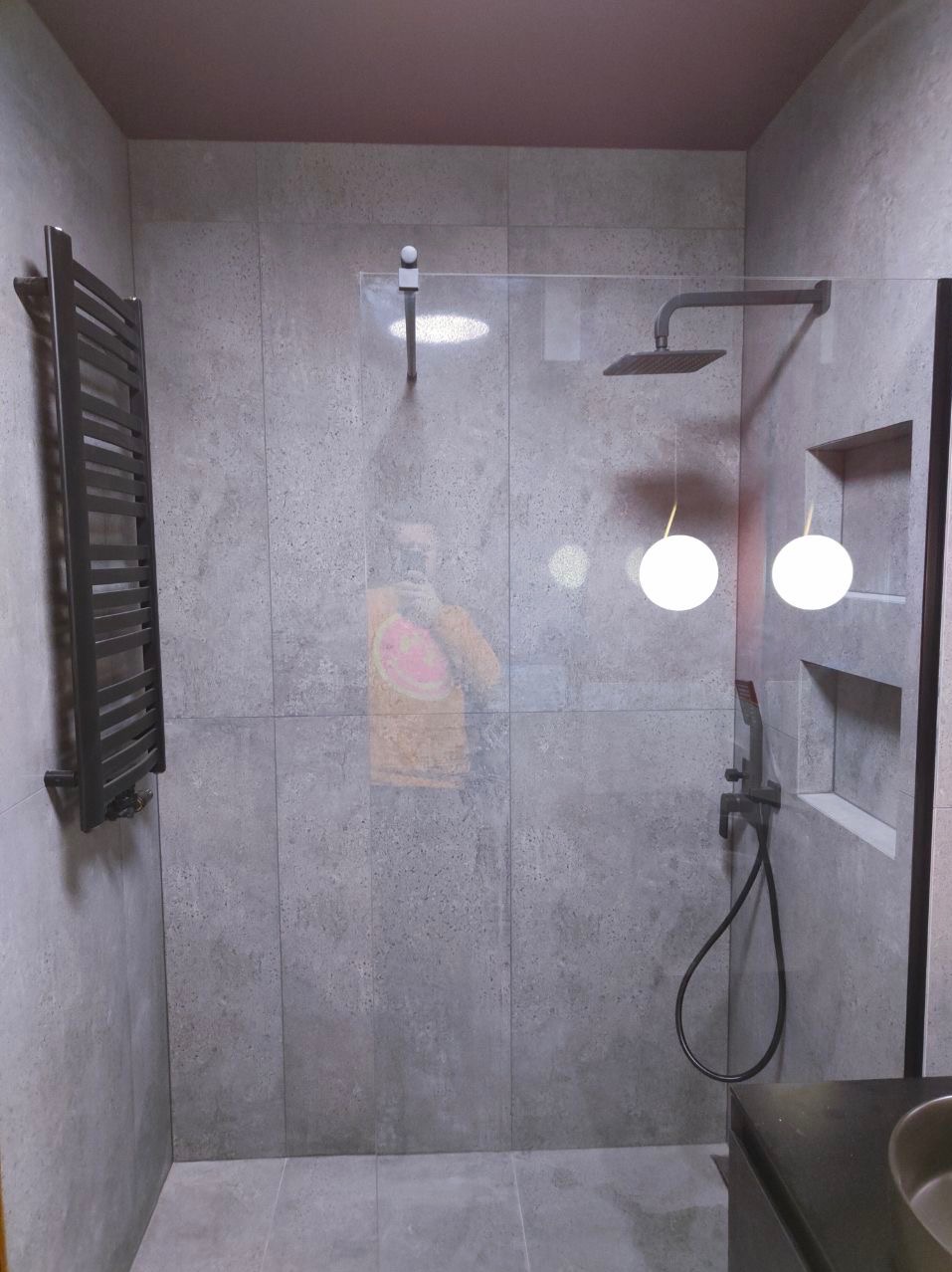 Nowoczesna kabina prysznicowa z deszczownicą i szklaną ścianką, szare płytki na ścianach, grzejnik drabinkowy po lewej, oświetlenie punktowe w niszach, widoczne odbicie osoby robiącej zdjęcie.