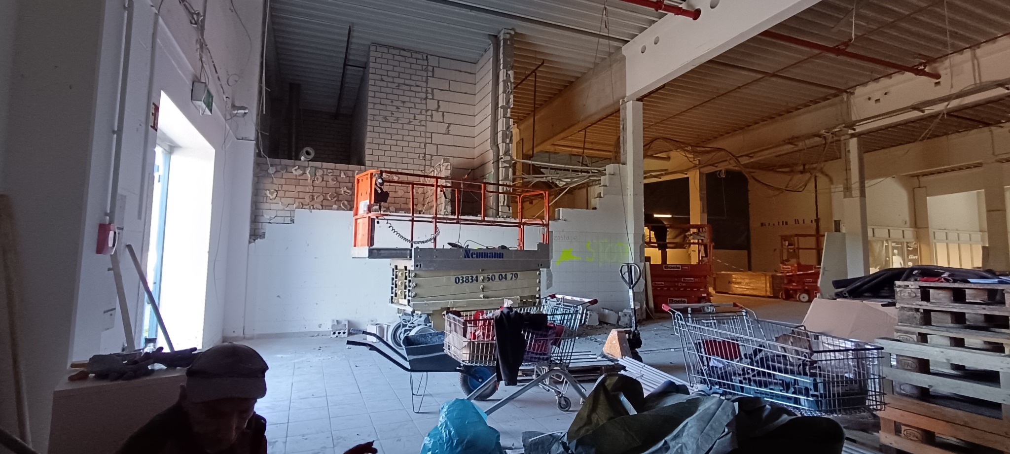 Wnętrze w trakcie wyburzania ścian z widocznym podnośnikiem nożycowym Neumann, resztkami muru z cegieł i wózkami sklepowymi wypełnionymi gruzem.