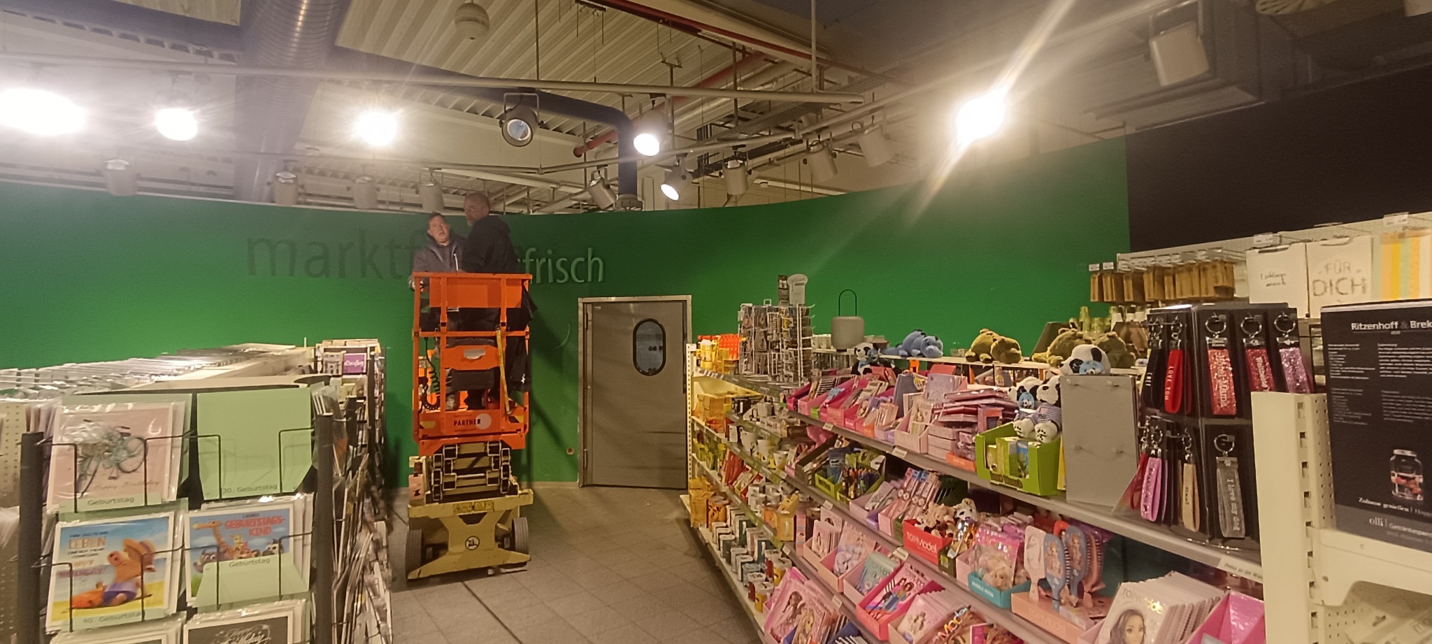Dwóch mężczyzn na podnośniku koszowym przed zieloną ścianą z napisem 'markt frisch' wewnątrz sklepu, prawdopodobnie podczas prac remontowych lub wykończeniowych.