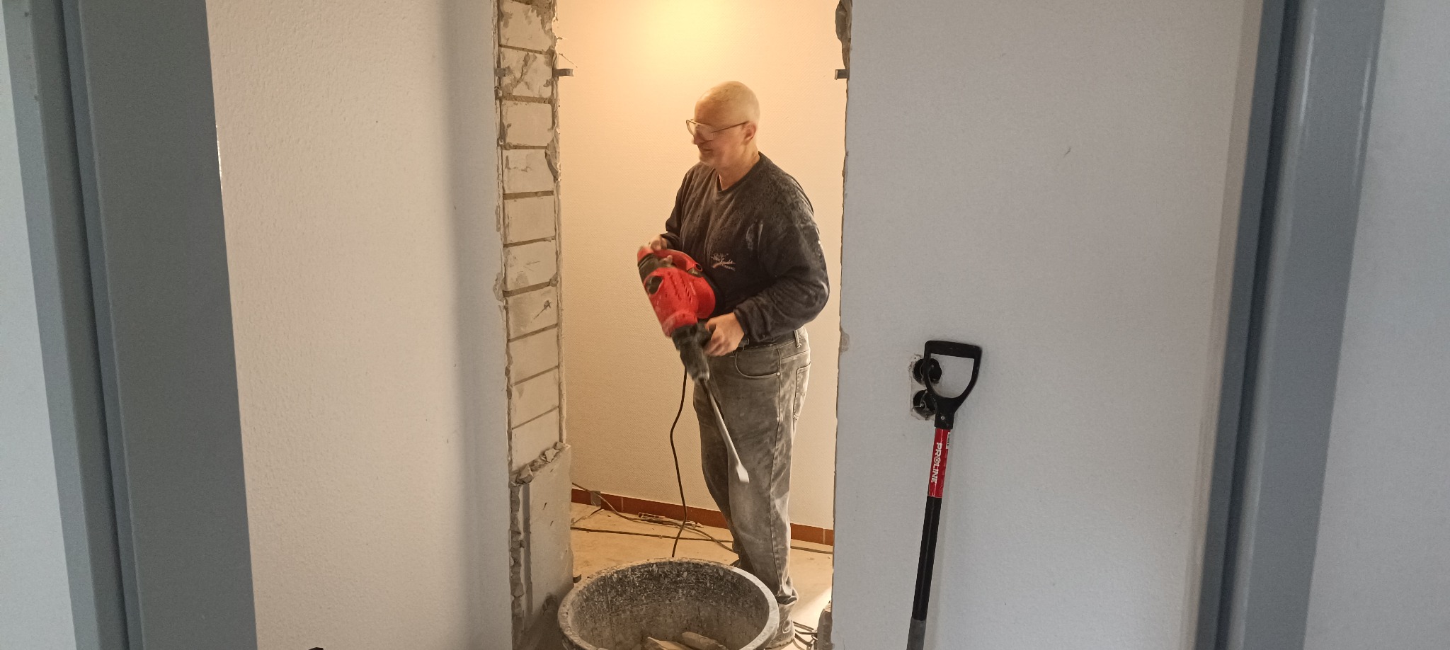 Mężczyzna w okularach używa czerwonego młota udarowego do wyburzenia fragmentu ściany z cegieł, widoczny kubeł z gruzem i łopata oparta o ścianę.