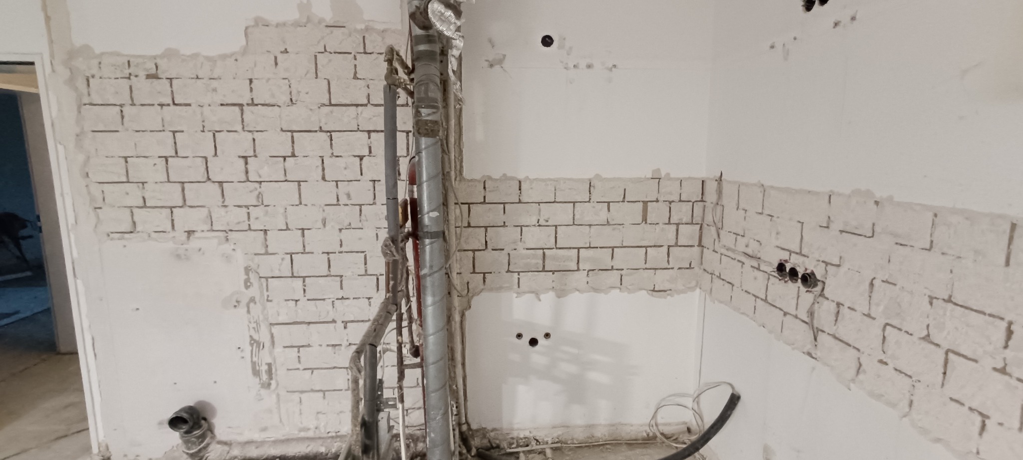 Fragment ściany w Trzebnicy po skuciu płytek, widoczne ślady po kleju w kształcie cegiełek, rury instalacyjne i otwory na gniazdka elektryczne.