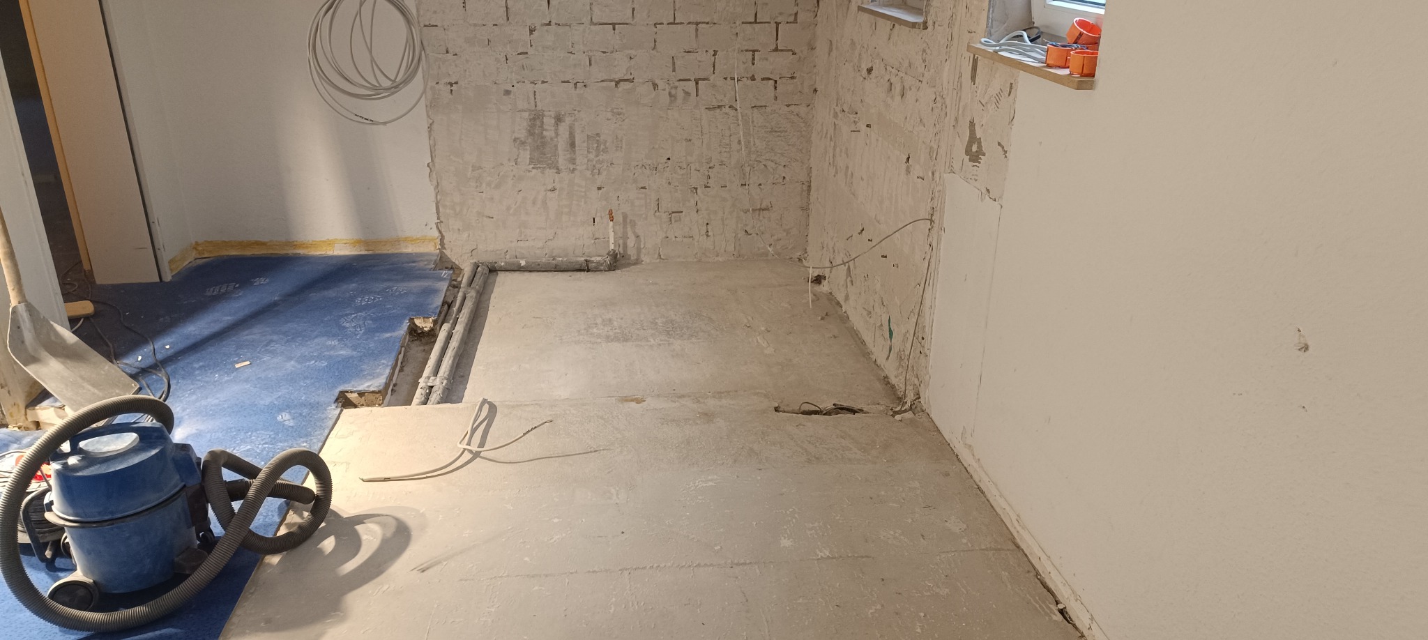 Wnętrze w trakcie prac remontowych: ogołocone cegły, fragment instalacji wodnej, kabel elektryczny wystający z podłogi, niebieska folia ochronna i odkurzacz przemysłowy.