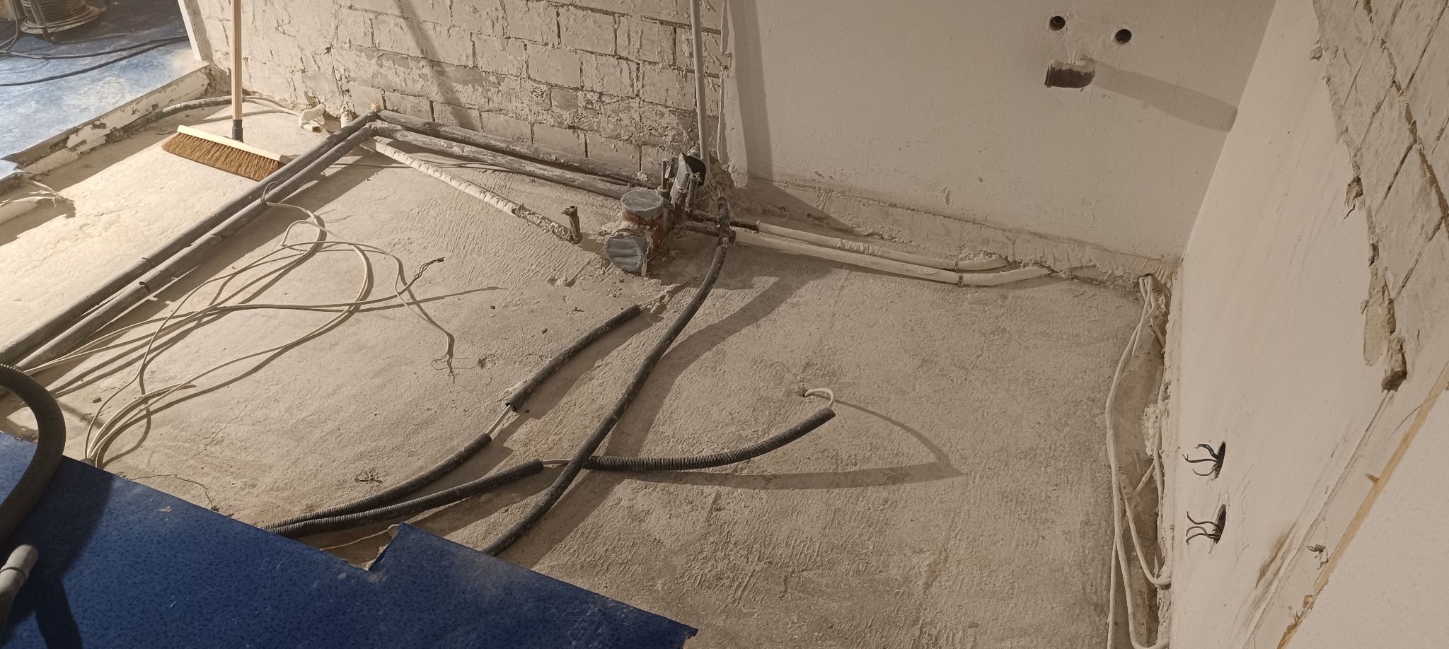 Wnętrze w trakcie remontu: odsłonięte rury, przewody elektryczne wystające ze ściany, resztki gruzu i pyłu na podłodze, widoczny fragment ściany z cegły i otwory w ścianie. Na podłodze leży szczotka.
