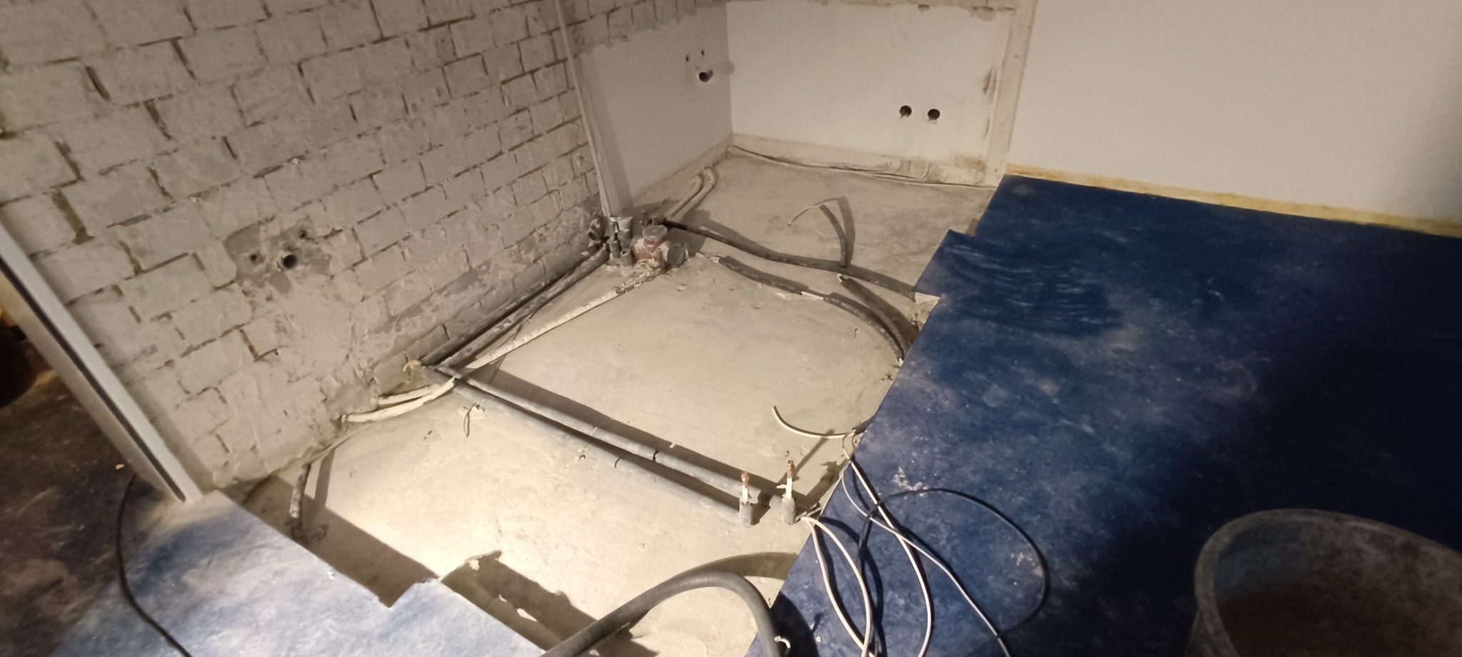 Fragment pomieszczenia w trakcie remontu: odsłonięta ceglana ściana, wystające rury i kable, częściowo zabezpieczona podłoga niebieską folią.