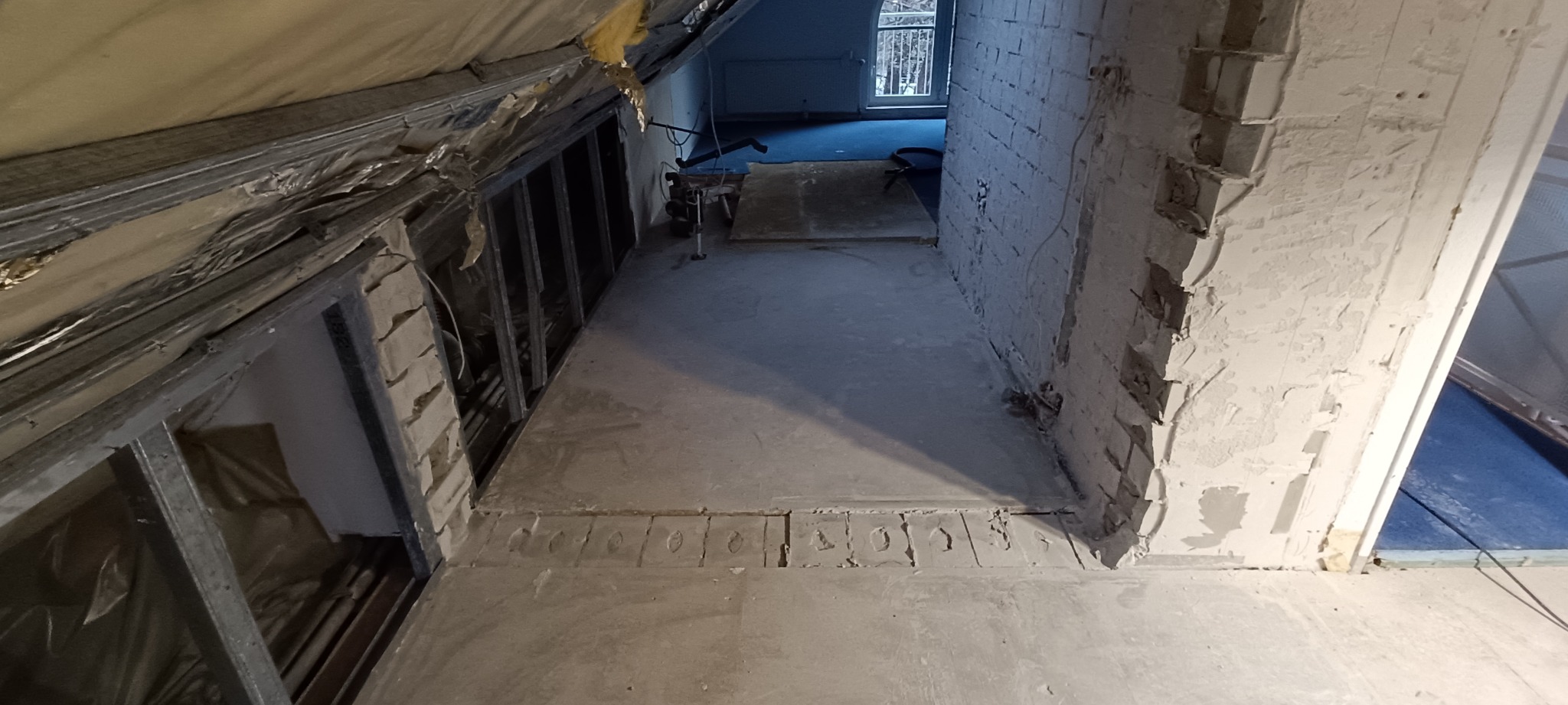 Wnętrze poddasza w trakcie wyburzania ściany działowej z odsłoniętą cegłą, widoczne profile metalowe konstrukcji, izolacja oraz fragment okna w tle.