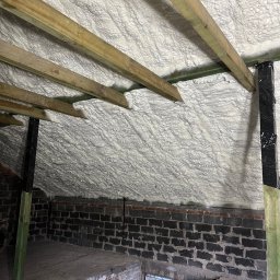 Hydrotherm - Poddasze po izolacji, widoczne drewniane krokwie i ściana z ciemnej cegły, biała izolacja szczelnie wypełnia przestrzeń pod dachem.