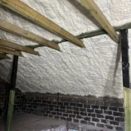 Poddasze po izolacji, widoczne drewniane krokwie i ściana z ciemnej cegły, biała izolacja szczelnie wypełnia przestrzeń pod dachem.