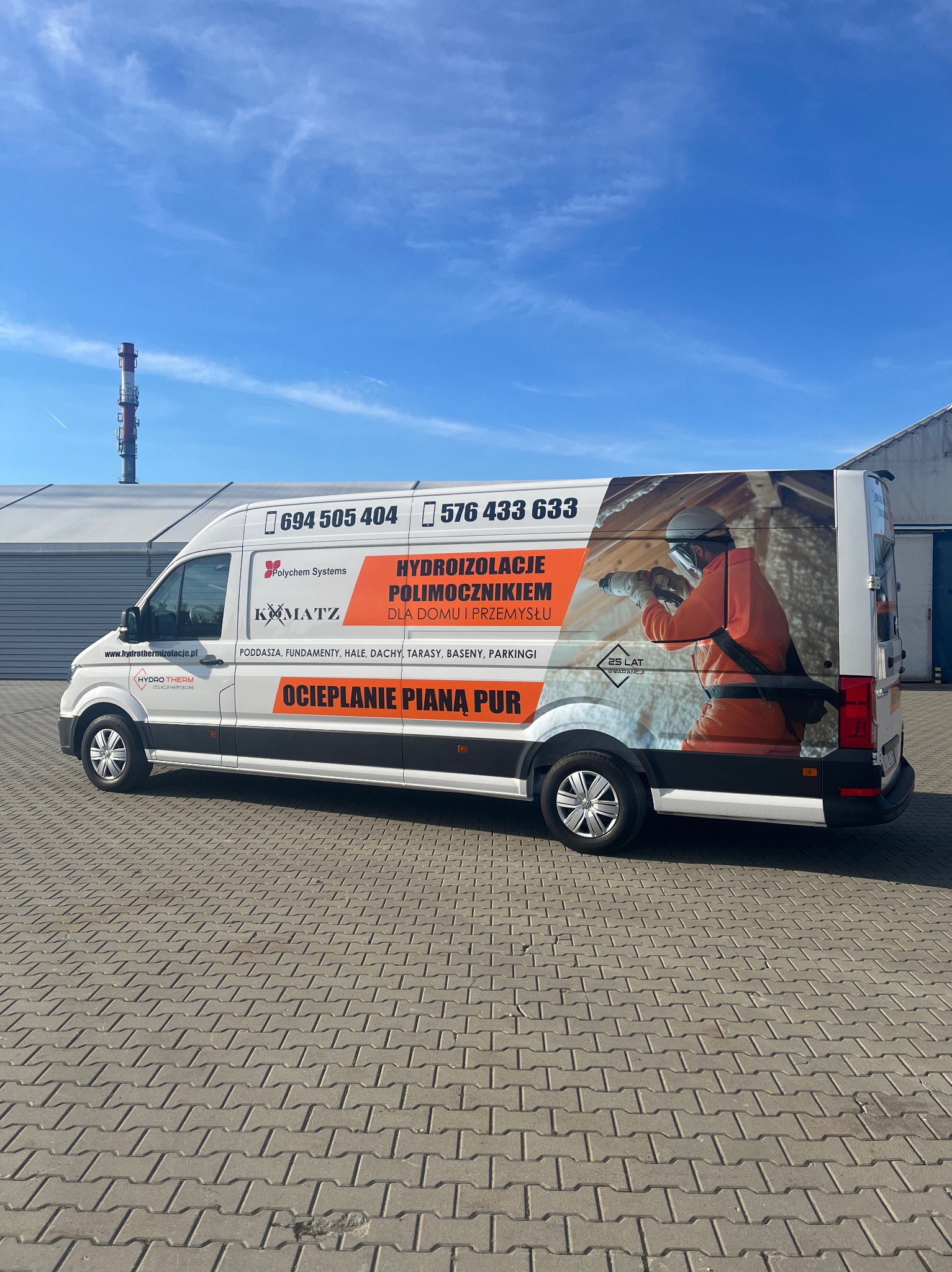 Biały samochód dostawczy z reklamą firmy oferującej hydroizolacje i ocieplanie, na tle błękitnego nieba i przemysłowego krajobrazu.