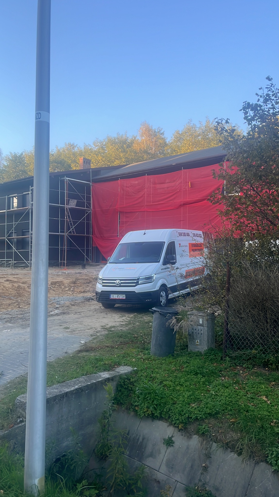 Budynek w trakcie prac ociepleniowych, osłonięty czerwoną plandeką, z rusztowaniami i zaparkowanym białym samochodem dostawczym z reklamą firmy.