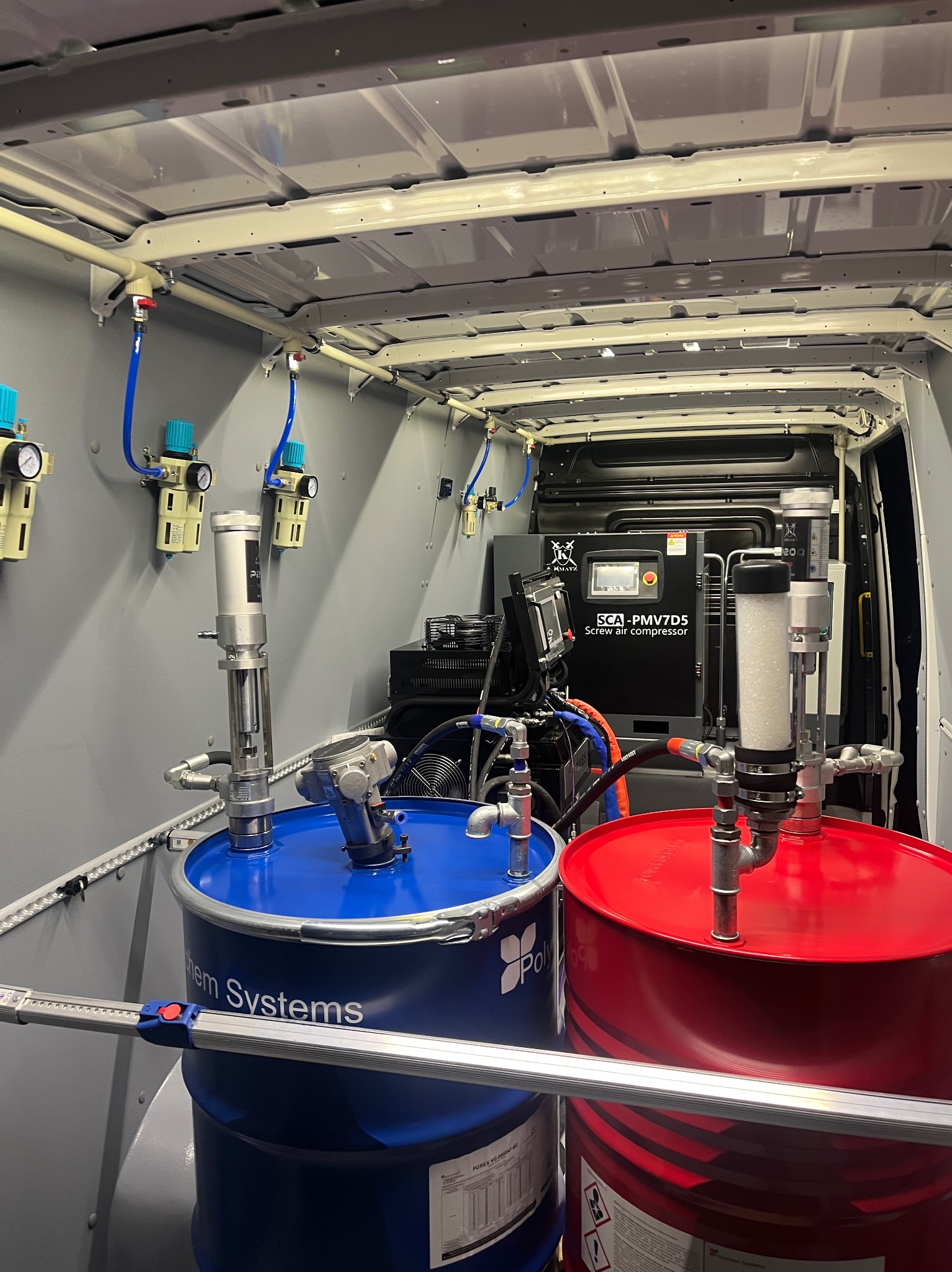 Wnętrze furgonetki z zamontowanym systemem do natrysku pianki PUR: dwa beczki (czerwona i niebieska) z pompami, kompresor śrubowy SCA-PMV7D5 i instalacja pneumatyczna.