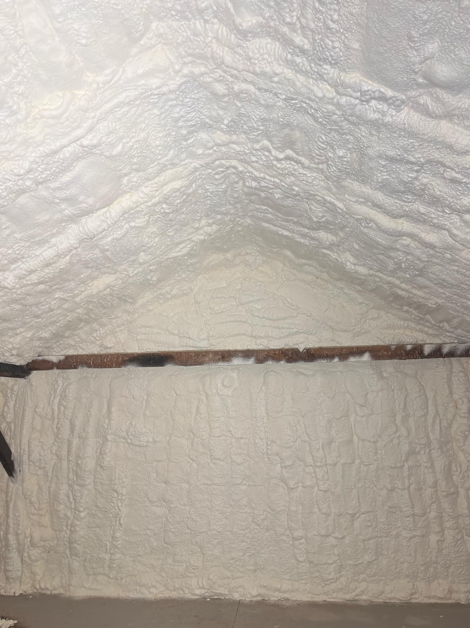 Ściana i skośny sufit poddasza pokryte grubą warstwą białej, nierównej izolacji natryskowej, widoczna drewniana belka konstrukcyjna.