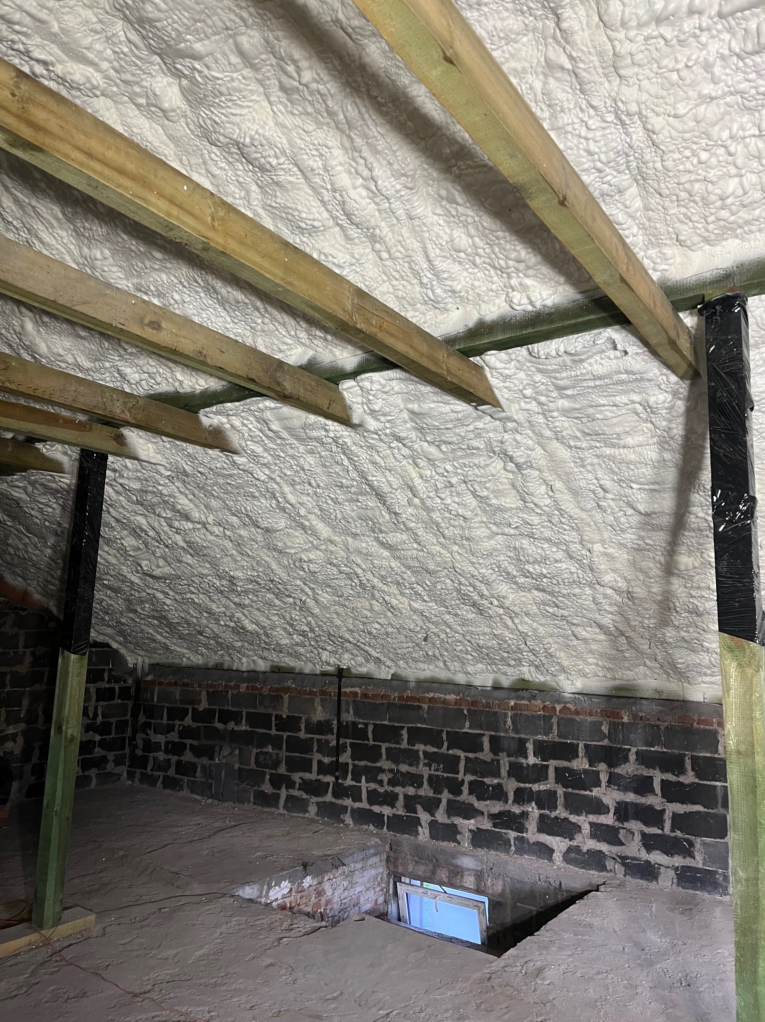 Poddasze po izolacji, widoczne drewniane krokwie i ściana z ciemnej cegły, biała izolacja szczelnie wypełnia przestrzeń pod dachem.