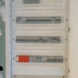 Otwarta skrzynka rozdzielcza z zainstalowanymi bezpiecznikami i zabezpieczeniami przeciwprzepięciowymi marki Simtec, widoczne diody sygnalizacyjne, tło stanowią surowe ściany w trakcie wykończenia.