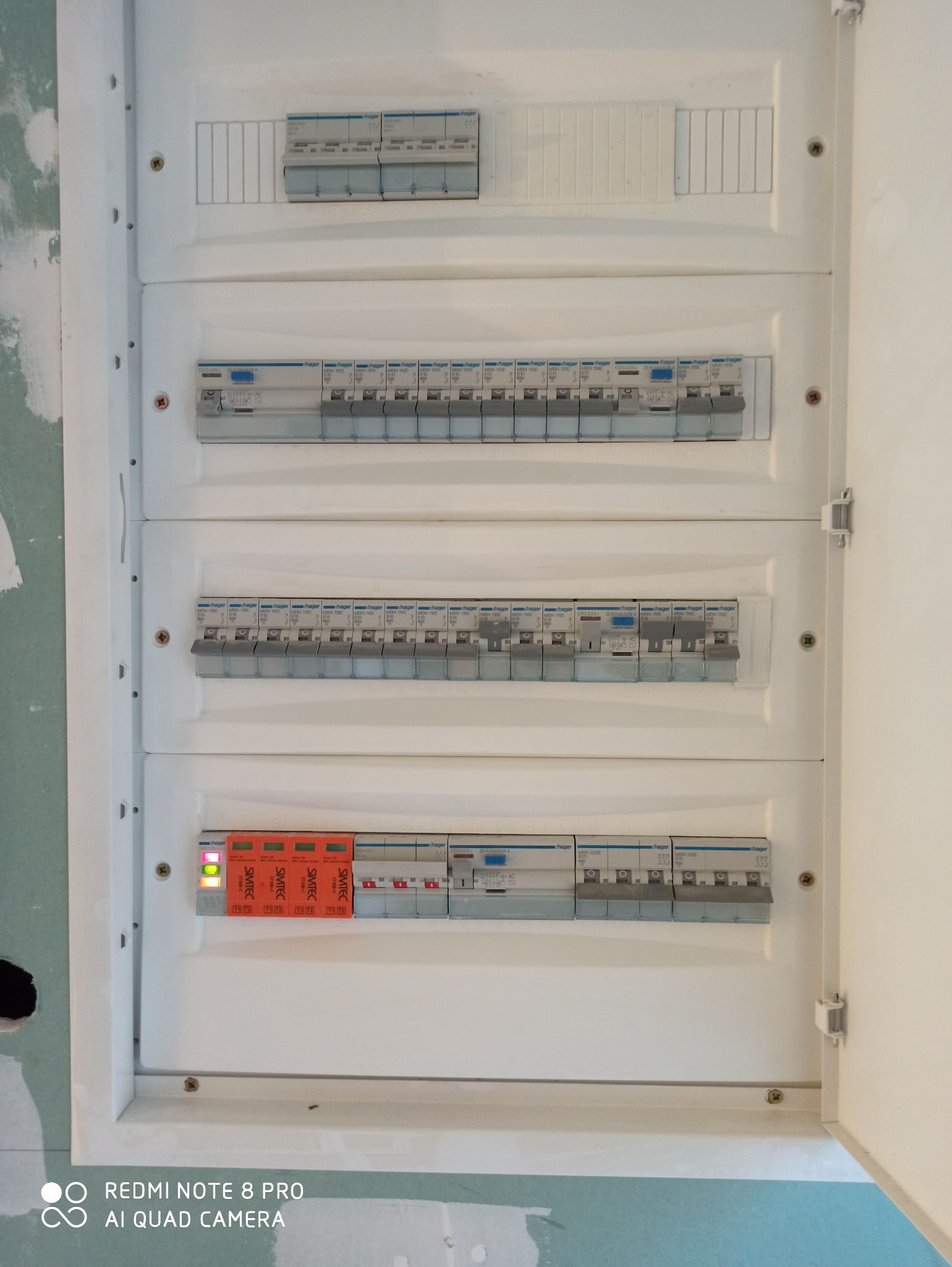 Otwarta skrzynka rozdzielcza z zainstalowanymi bezpiecznikami i zabezpieczeniami przeciwprzepięciowymi marki Simtec, widoczne diody sygnalizacyjne, tło stanowią surowe ściany w trakcie wykończenia.