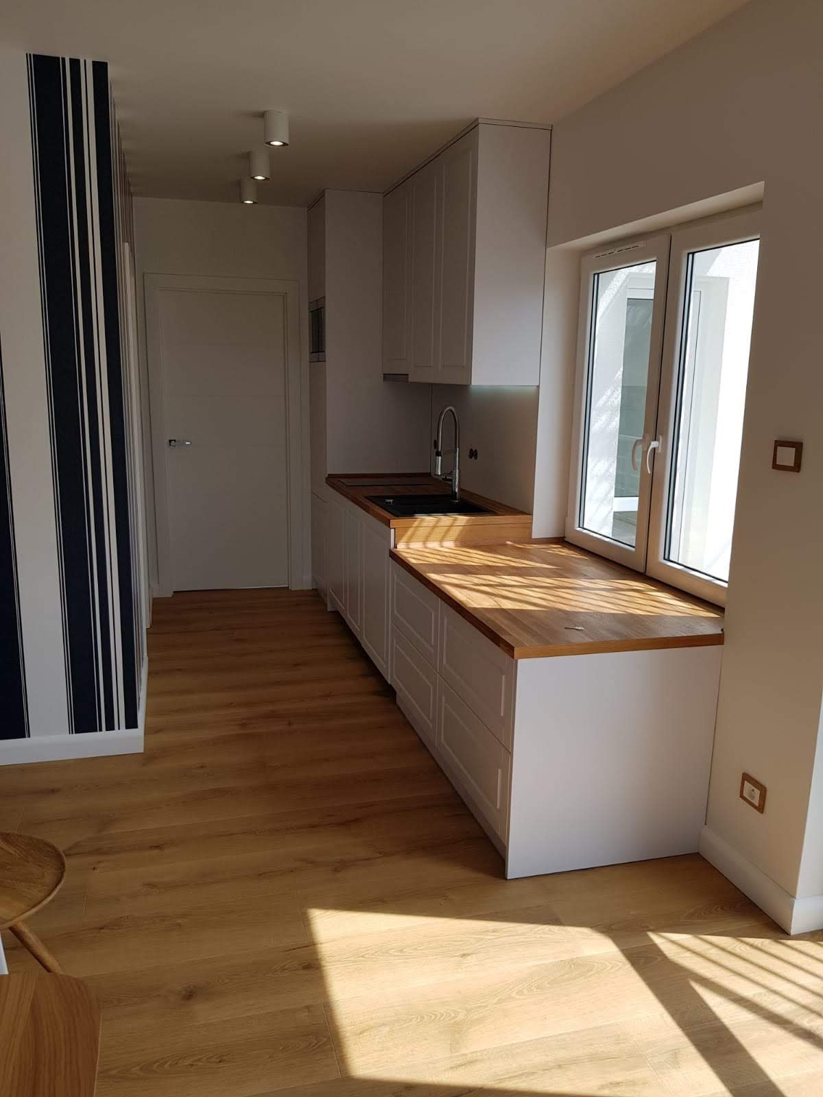 Jasna kuchnia z drewnianym blatem, białymi szafkami i oknem. Na ścianie dekoracyjne pasy. Podłoga z paneli. Oświetlenie sufitowe punktowe. Minimalistyczny design.