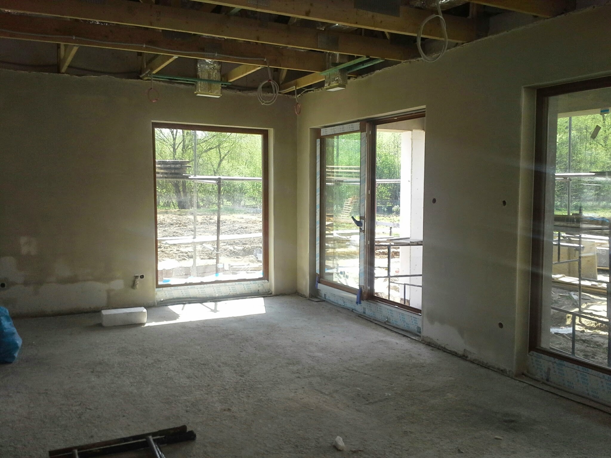 Surowy stan wnętrza z oknami i drewnianą konstrukcją sufitu. Ściany przygotowane do dalszych prac wykończeniowych, widoczne instalacje pod sufitem.