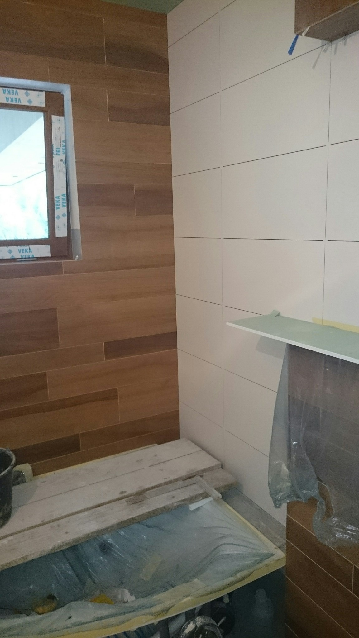 Ukończona ściana z białych płytek i fragment ściany z płytek imitujących drewno w łazience. Widoczne zabezpieczone elementy podczas prac wykończeniowych.