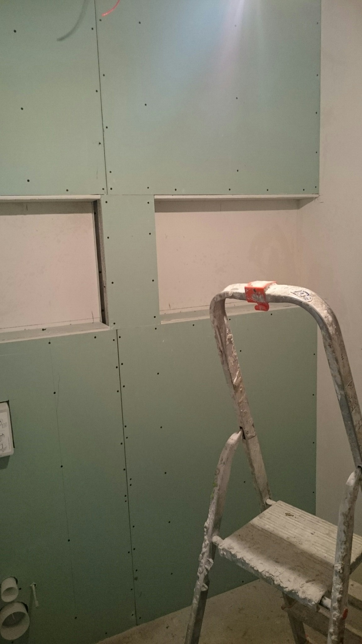 Ściana z zielonych płyt kartonowo-gipsowych z otworami, widoczna drabina malarska. Etap prac wykończeniowych w pomieszczeniu.