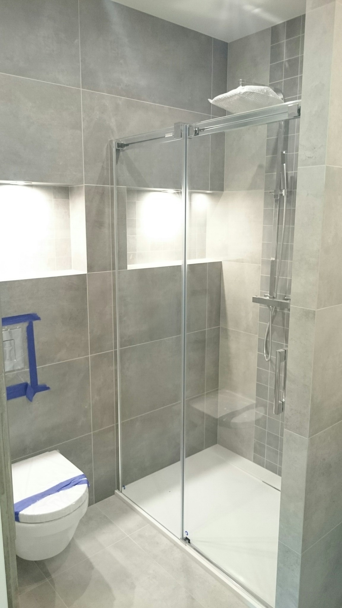 Nowoczesna łazienka z szarą glazurą, kabiną prysznicową typu walk-in i podświetlanymi niszami. Uwagę zwraca toaleta z niebieskim zabezpieczeniem.