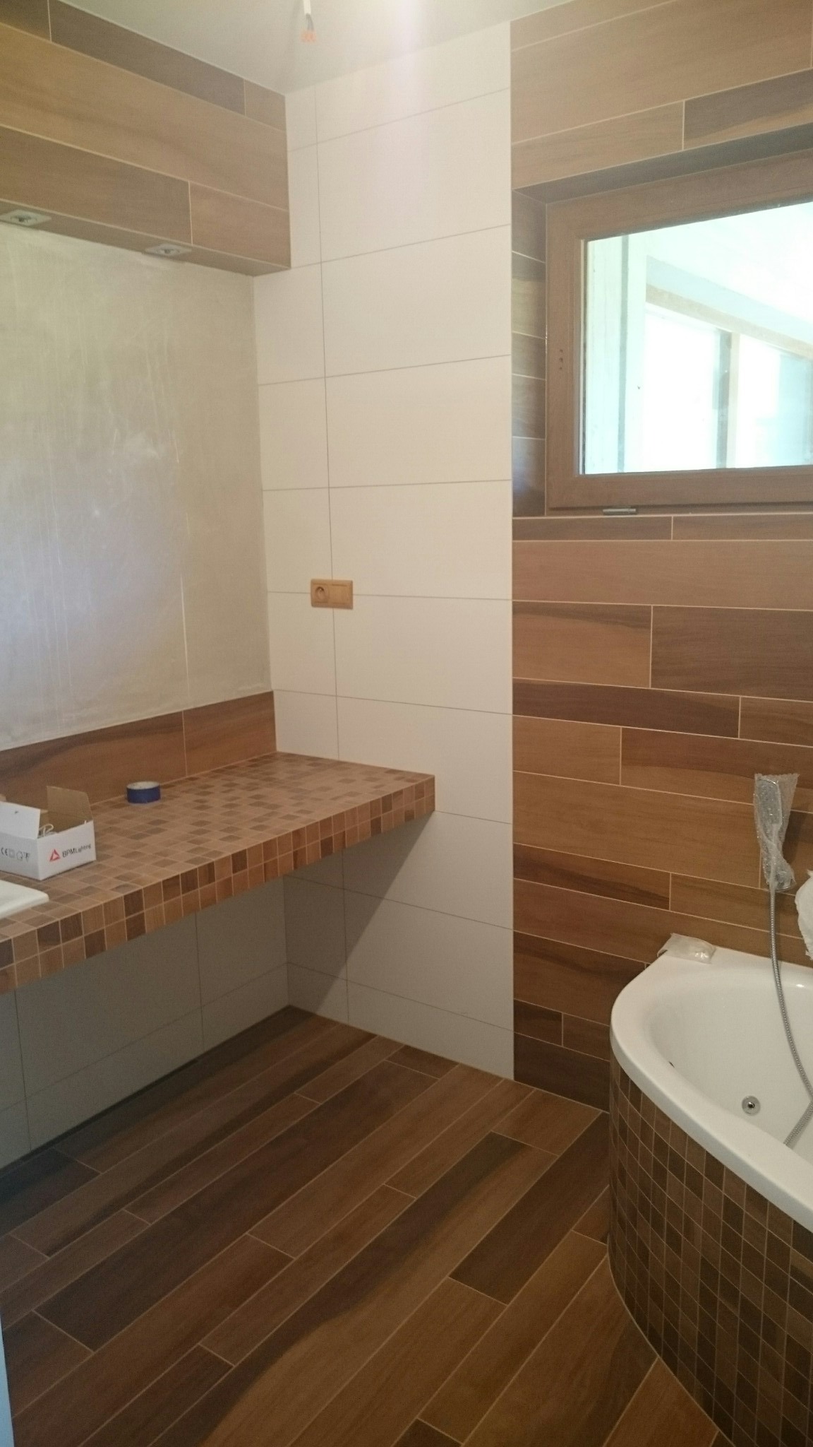 Wykończona łazienka z brązowymi płytkami imitującymi drewno na podłodze i ścianach, białe płytki na ścianie, zabudowa umywalki z mozaiki, wanna z hydromasażem.