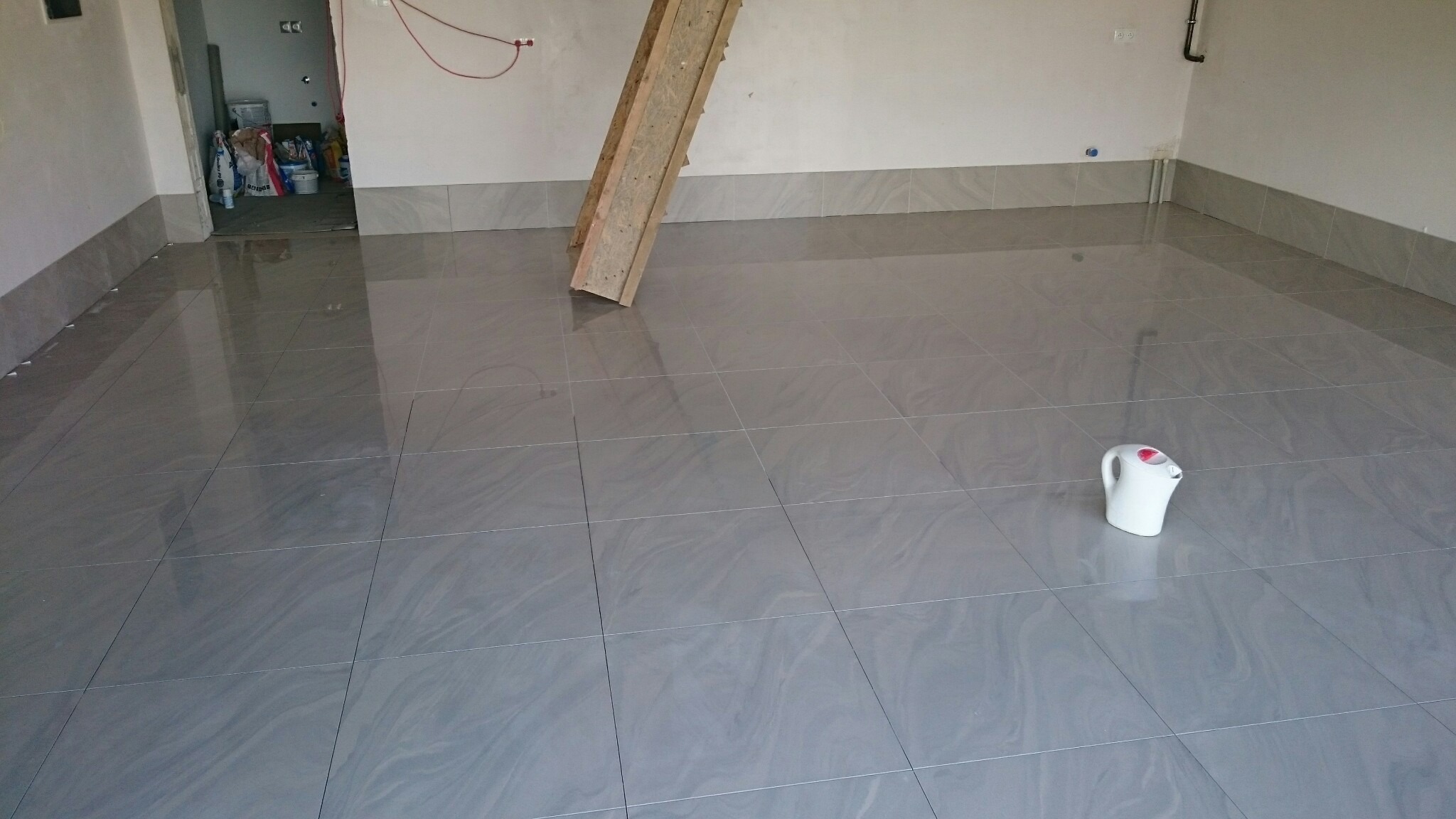 Położona, błyszcząca, szara podłoga z płytek. W tle widoczne schody drewniane i wejście do innego pomieszczenia. Na podłodze stoi biały czajnik.