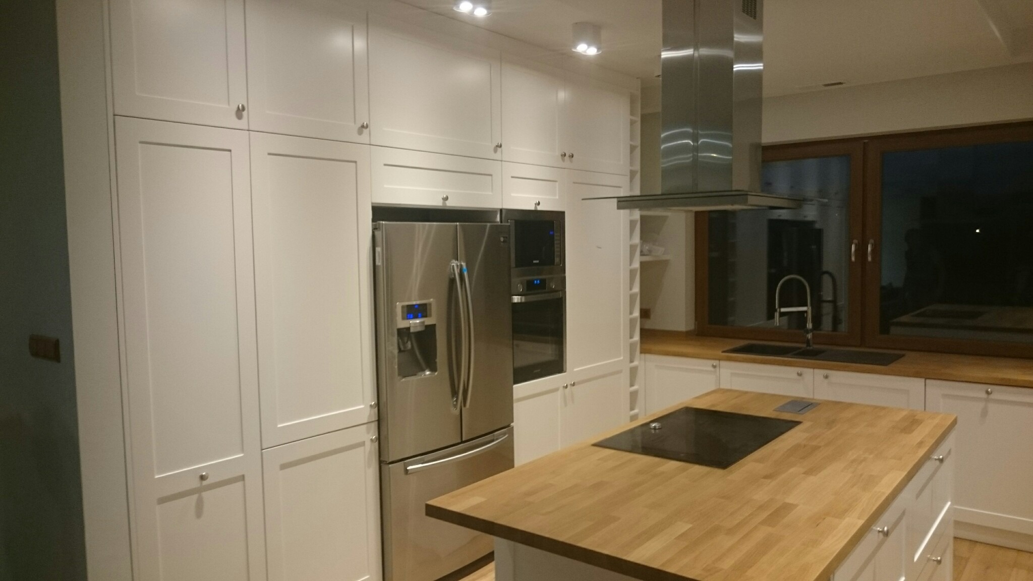 Nowoczesna kuchnia z białymi szafkami, drewnianym blatem wyspy i stalową lodówką. Minimalistyczny design, oświetlenie punktowe, okno z widokiem na zewnątrz.