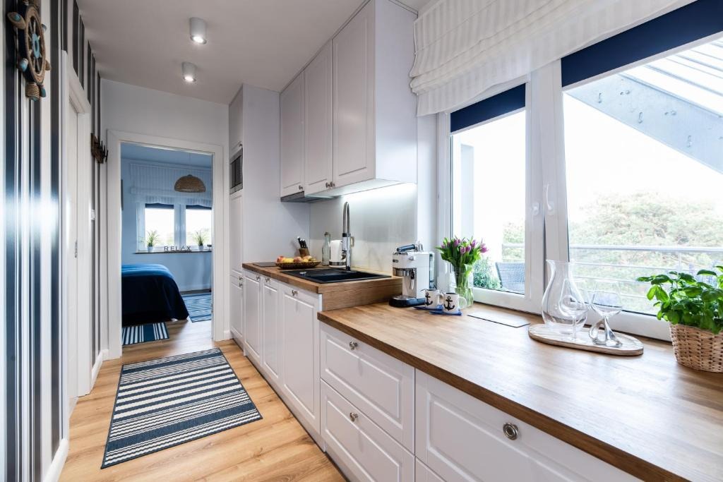 Jasna kuchnia w stylu marynistycznym z drewnianym blatem, białymi szafkami i widokiem na zieleń za oknem. W tle sypialnia z niebieskimi akcentami.