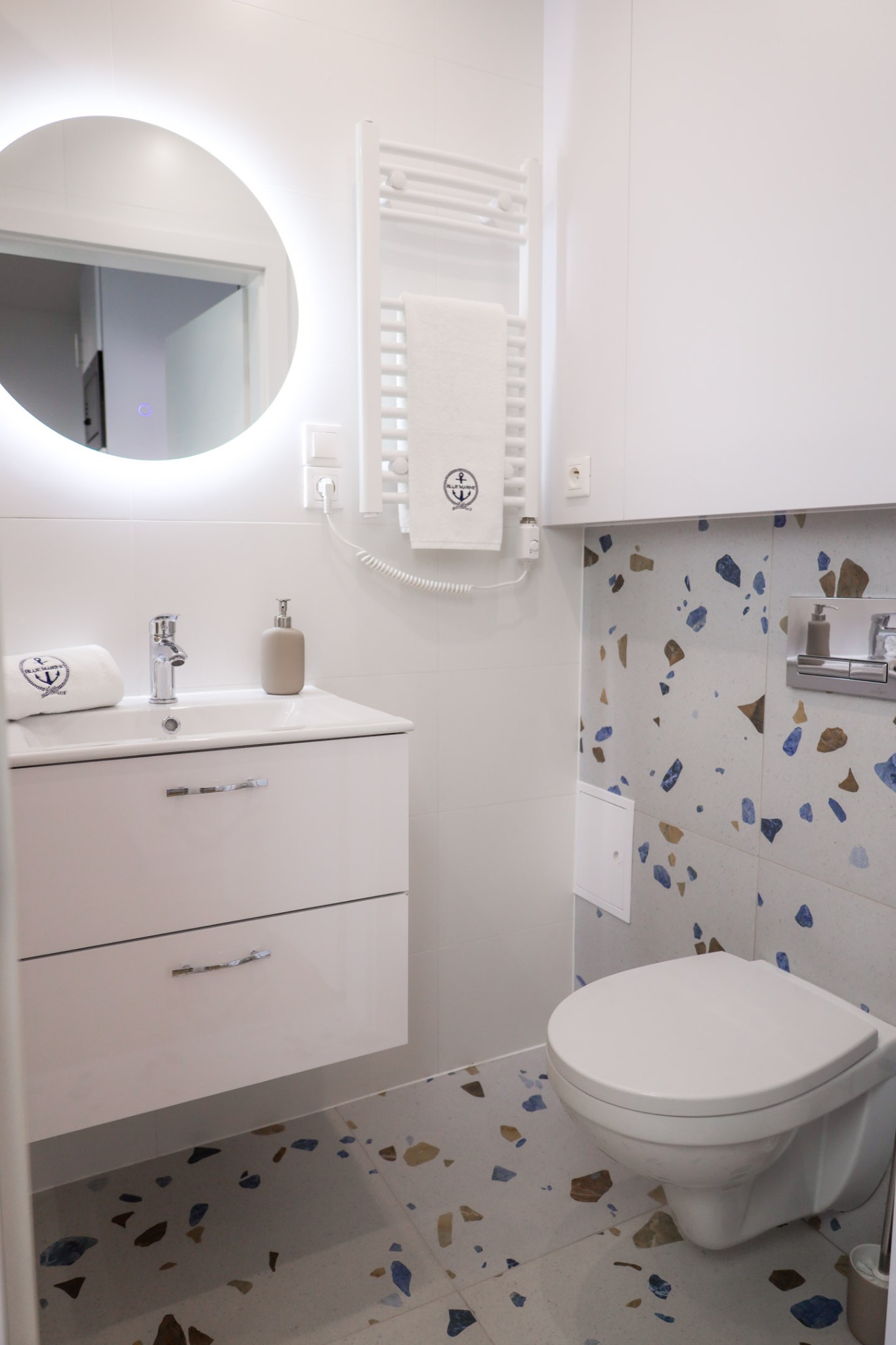 Biała łazienka z okrągłym lustrem z podświetleniem LED, umywalką z szafką i toaletą. Na ścianie i podłodze płytki w stylu lastryko z niebieskimi i brązowymi elementami.