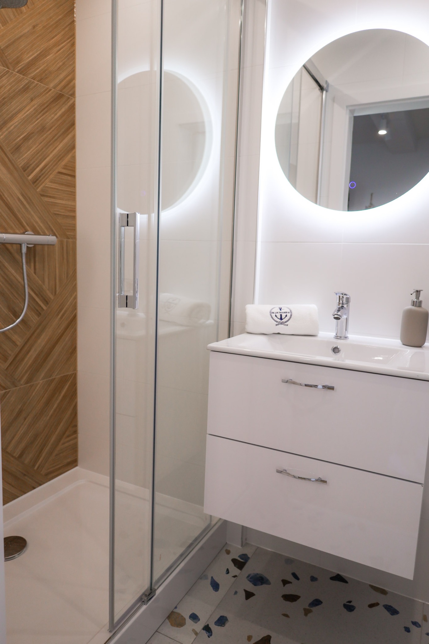 Nowoczesna łazienka z kabiną prysznicową, umywalką z szafką i okrągłym lustrem z podświetleniem LED. Uwagę przykuwają drewniane panele na ścianie prysznica i mozaika na podłodze.