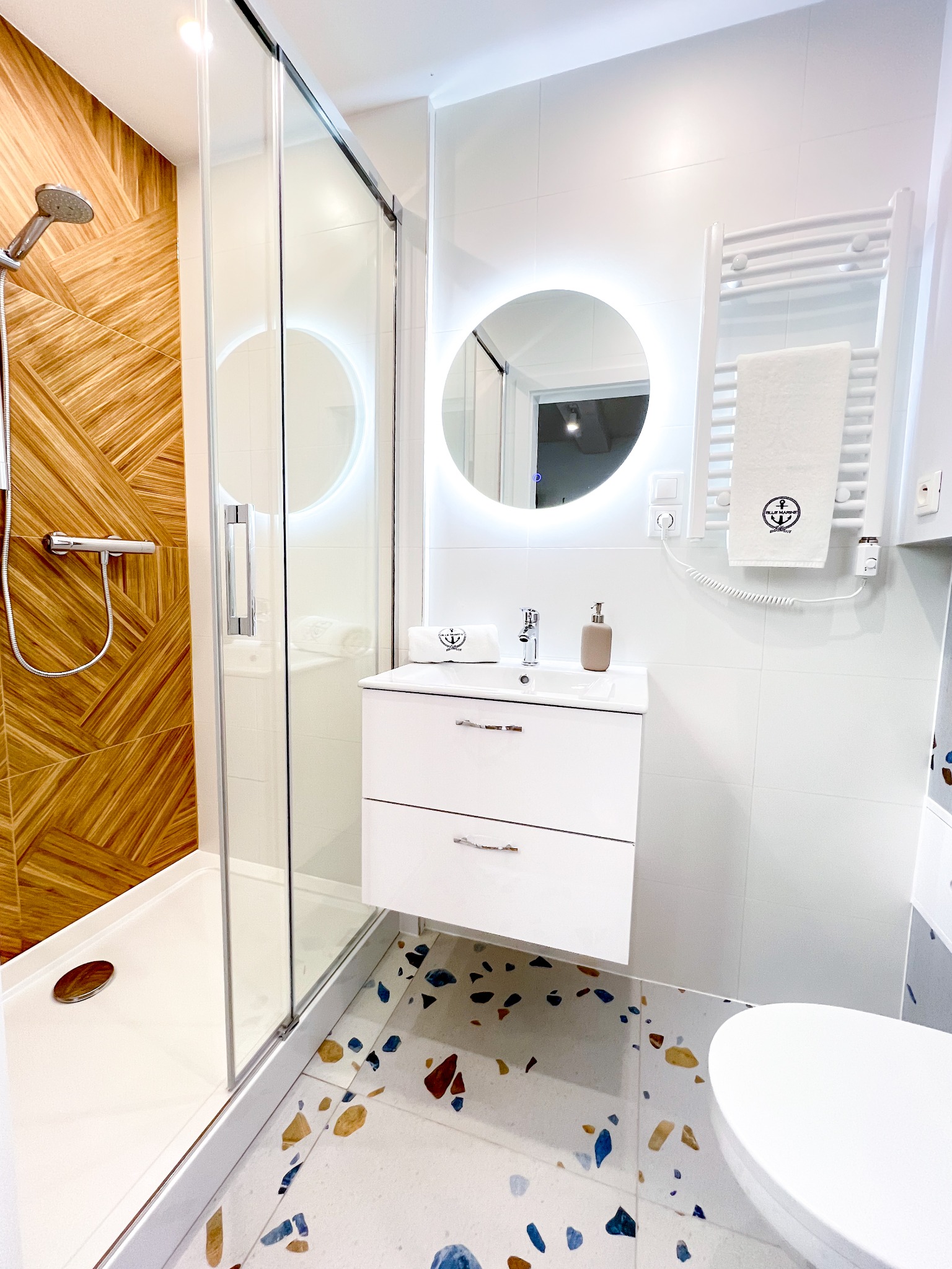 Nowoczesna łazienka z kabiną prysznicową wykończoną drewnopodobnymi płytkami, okrągłym lustrem z podświetleniem i mozaikową podłogą.