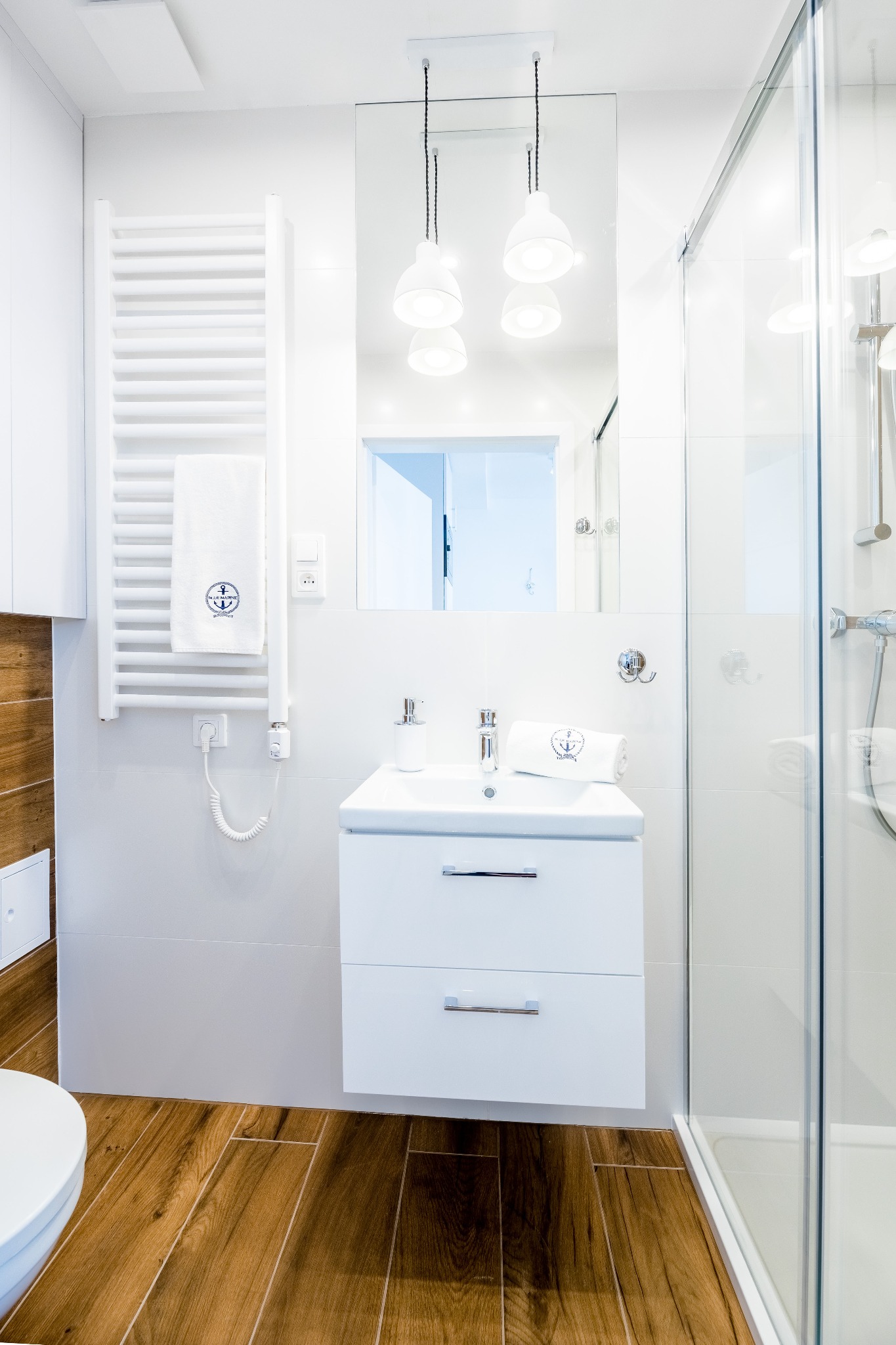 Jasna łazienka z drewnianą podłogą i białymi meblami. Kabina prysznicowa ze szkła, biała umywalka z szafką i lustro z lampami. Grzejnik drabinkowy z ręcznikiem.