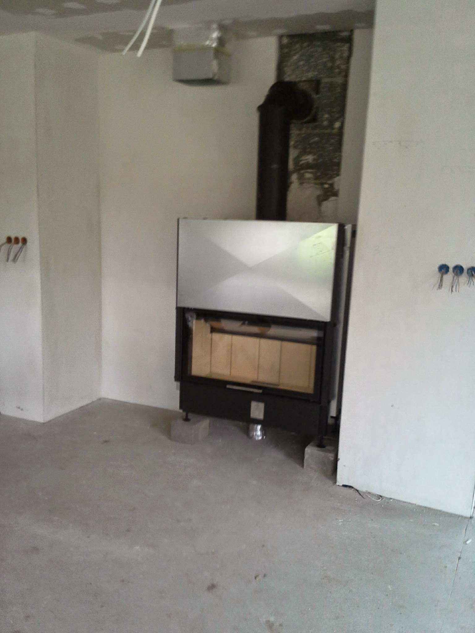 Wnętrze w trakcie prac wykończeniowych z kominkiem. Widoczne przewody elektryczne i fragmenty instalacji kominowej. Ściany przygotowane do dalszych prac.