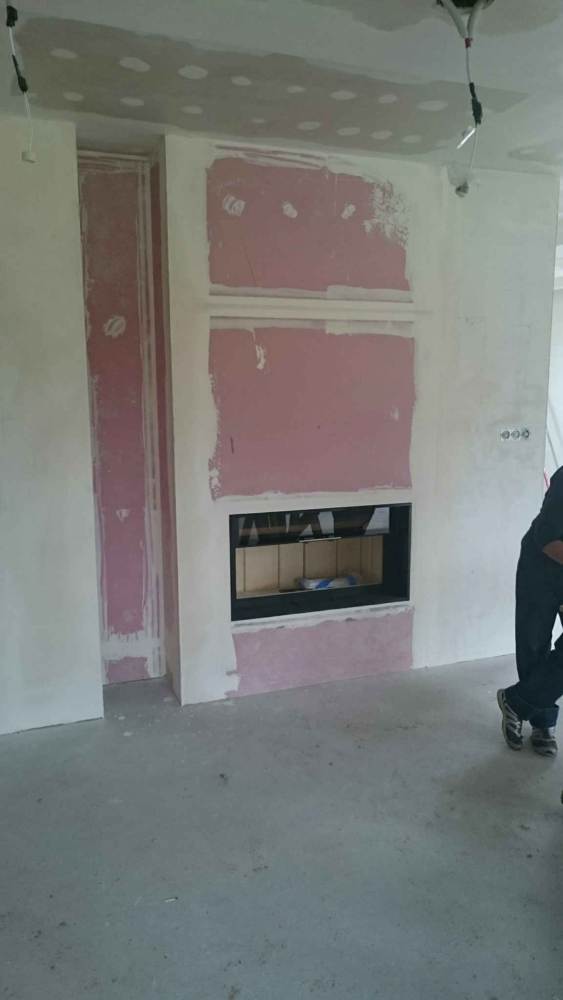 Wnętrze w trakcie prac wykończeniowych: zabudowa kominka z płyt kartonowo-gipsowych, ściany częściowo pokryte różową warstwą, widoczne przewody elektryczne.