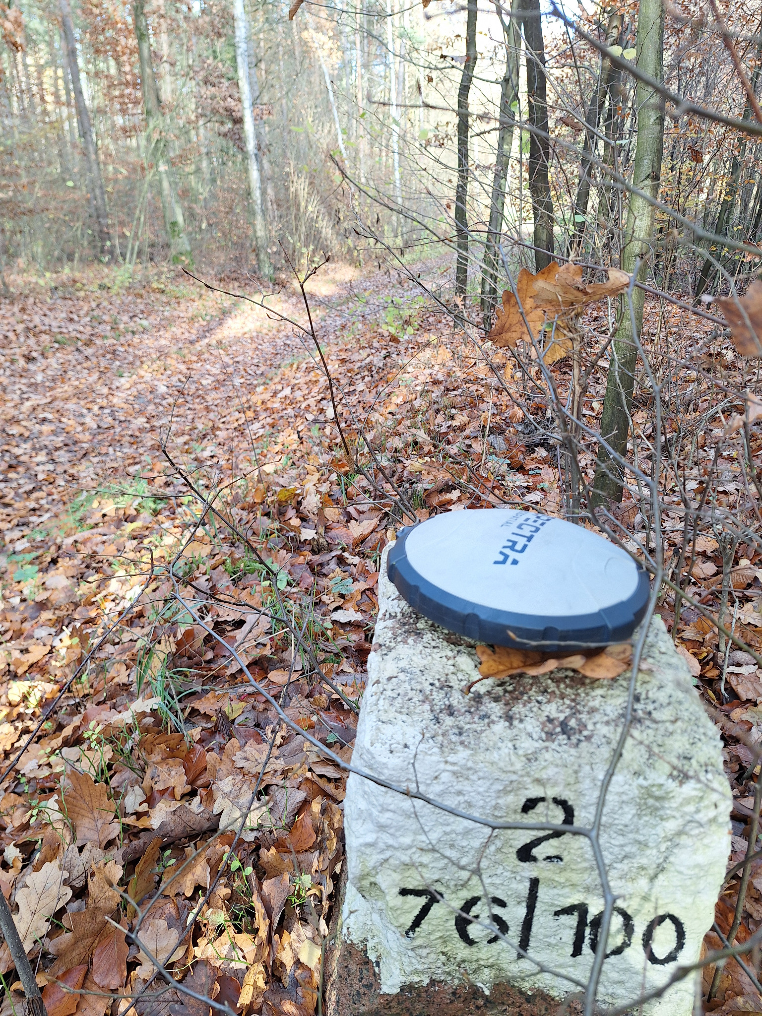 Graniczny słup z numerem 2 76/100 w jesiennym lesie, na nim leży odbiornik GPS marki Spectra.