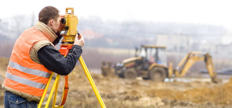 Pracownik w pomarańczowej kamizelce mierzy teren teodolitem na żółtym statywie; w tle rozmyta koparka i zabudowa.