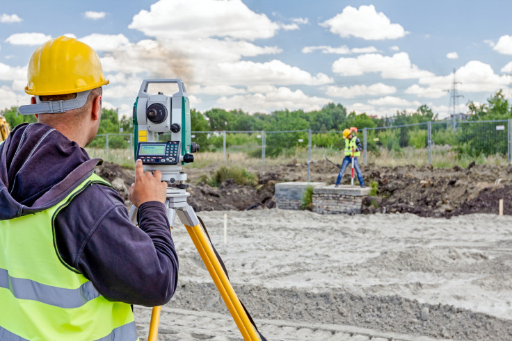 Pracownik w żółtym kasku obsługuje teodolit na budowie, w tle drugi pracownik wbija palik.