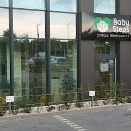 Centrum Terapii Dziecka BABY STEPS Częstochowa - fizjoterapia - logopeda dziecięcy - psycholog dziecięcy - Budynek z dużymi oknami i szyldem 'Baby Steps Centrum Terapii Dziecka' z zielonym logo w kształcie serca i dłoni. Przed budynkiem widoczne rośliny i oznakowanie parkingowe.