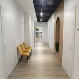 Centrum Terapii Dziecka BABY STEPS Częstochowa - fizjoterapia - logopeda dziecięcy - psycholog dziecięcy - Jasny, nowoczesny korytarz w placówce medycznej z rzędem trzech żółtych krzeseł pod ścianą. Na suficie widoczne podłużne lampy. Po prawej stronie ściana z owalnym, dekoracyjnym wcięciem.