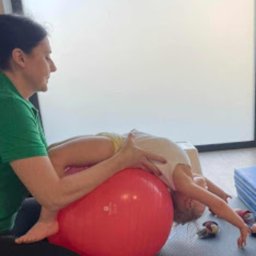 Centrum Terapii Dziecka BABY STEPS Częstochowa - fizjoterapia - logopeda dziecięcy - psycholog dziecięcy - Dziecko leżące na czerwonej piłce rehabilitacyjnej, podtrzymywane przez kobietę w zielonej koszulce podczas ćwiczeń w jasnym pomieszczeniu.