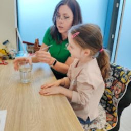 Centrum Terapii Dziecka BABY STEPS Częstochowa - fizjoterapia - logopeda dziecięcy - psycholog dziecięcy - Kobieta w zielonej koszulce pokazuje dziewczynce z różową spinką do włosów ćwiczenie z wodą i patyczkiem przy stole z jasnego drewna, na krześle z zabawnym wzorem.