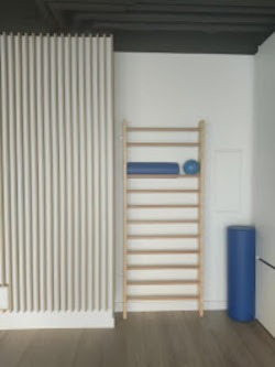 Wnętrze gabinetu z drewnianą drabinką gimnastyczną, na której leży niebieski wałek i piłka. Obok drabinki stoi niebieski wałek do ćwiczeń. Ściana pomalowana na biało, a podłoga jest drewniana.