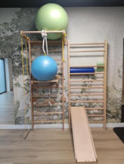 Drewniane drabinki gimnastyczne z linową siatką i piłkami rehabilitacyjnymi w jasnym pomieszczeniu z tapetą w motywy leśne.