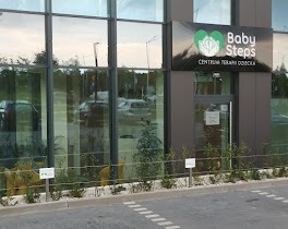 Budynek z dużymi oknami i szyldem 'Baby Steps Centrum Terapii Dziecka' z zielonym logo w kształcie serca i dłoni. Przed budynkiem widoczne rośliny i oznakowanie parkingowe.