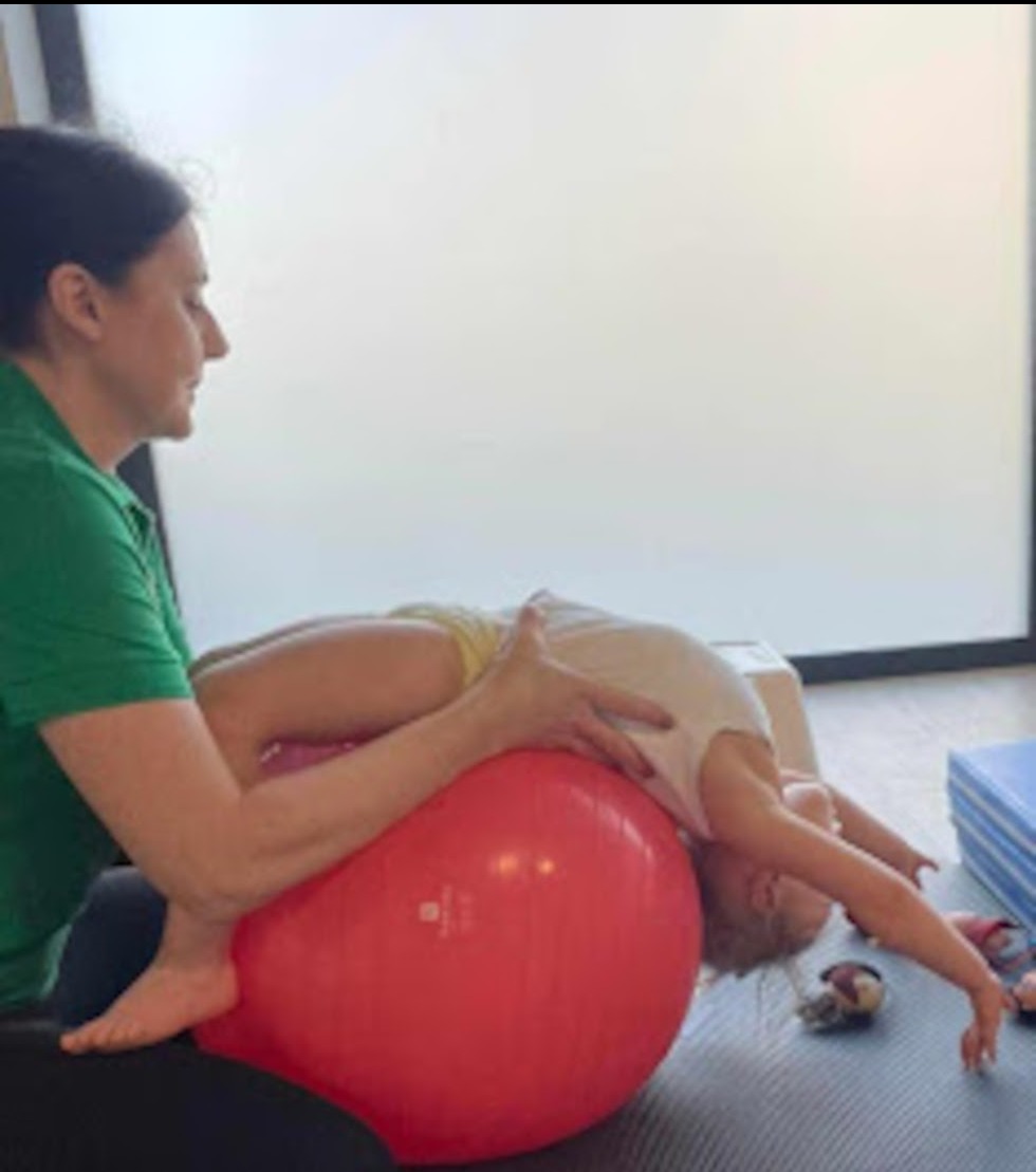 Dziecko leżące na czerwonej piłce rehabilitacyjnej, podtrzymywane przez kobietę w zielonej koszulce podczas ćwiczeń w jasnym pomieszczeniu.