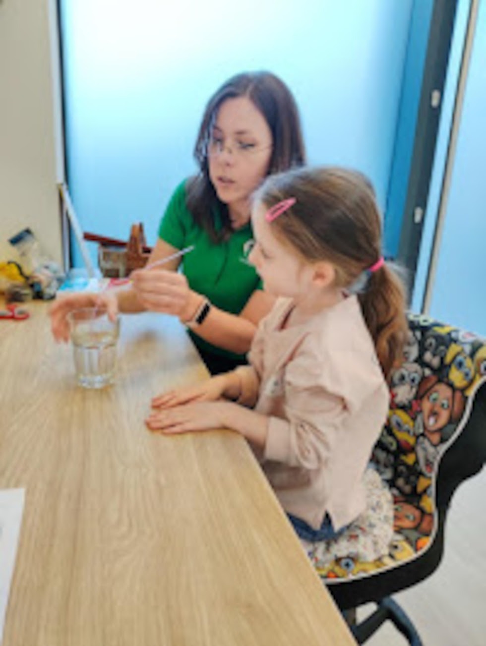 Kobieta w zielonej koszulce pokazuje dziewczynce z różową spinką do włosów ćwiczenie z wodą i patyczkiem przy stole z jasnego drewna, na krześle z zabawnym wzorem.